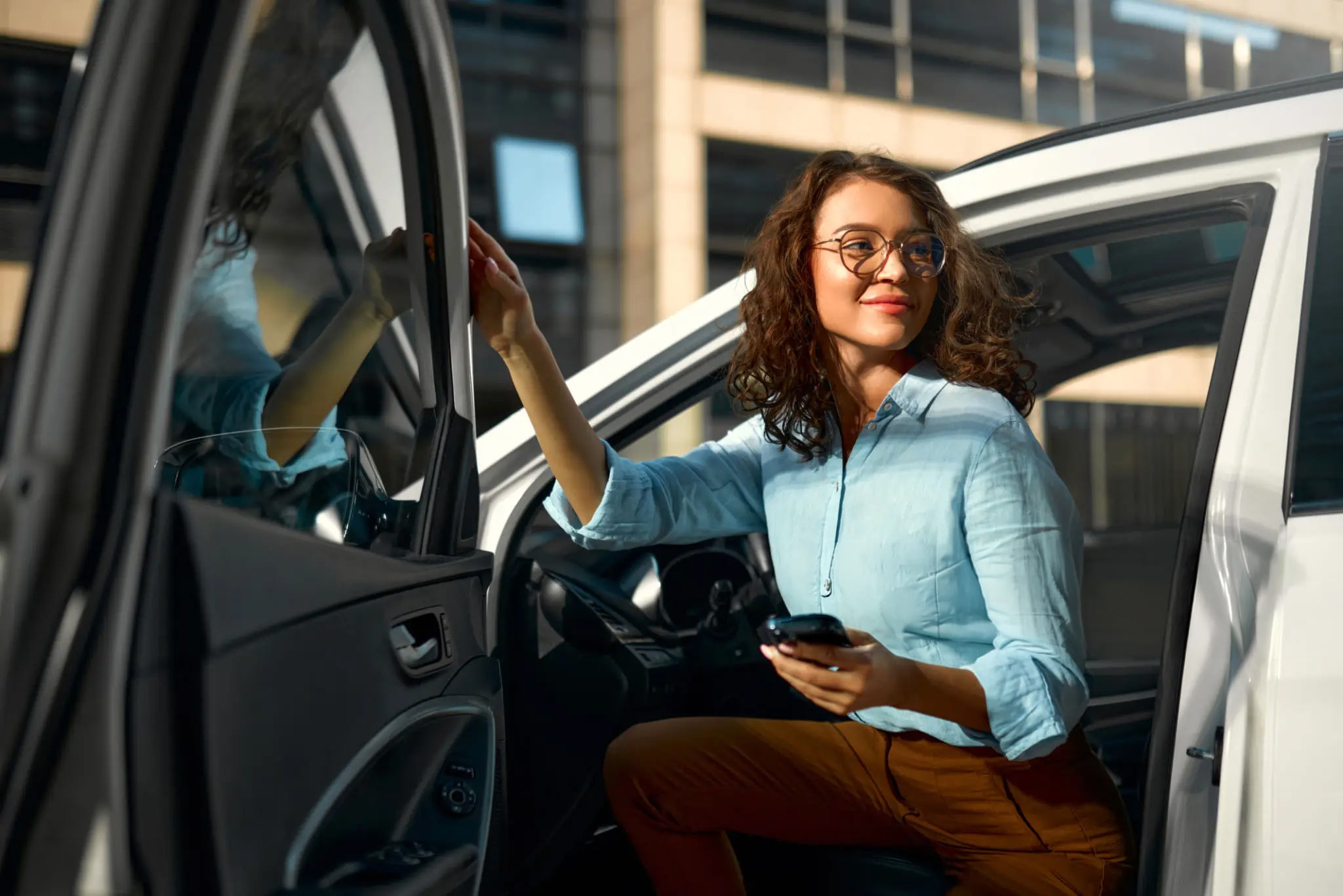 Frau mit Brille und blauem Hemd steigt mit Smartphone in der Hand in ein weißes Auto ein.